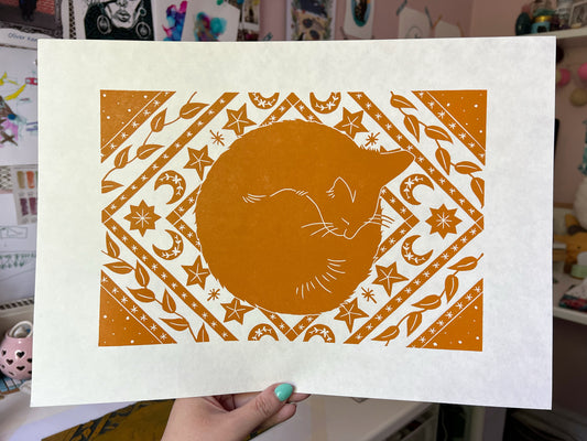 An A3 orange lino print of a cat napping on a celestial rug