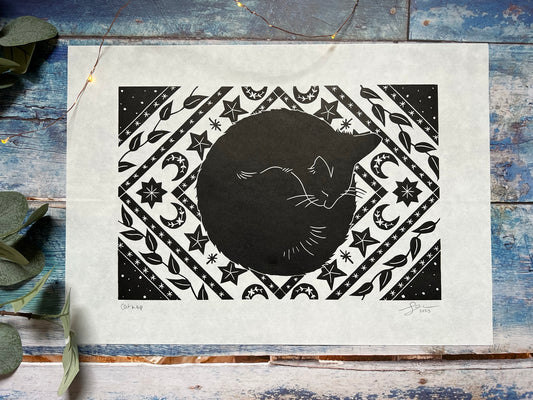 An A3 lino print of a black cat napping on a rug