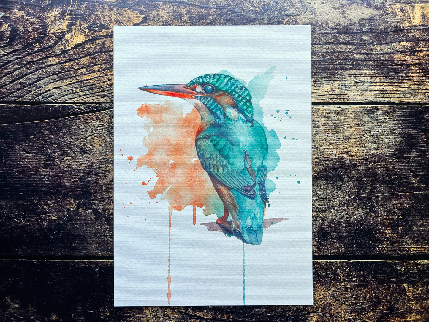 A photograph of a print of a watercolour kingfisher. It's painted in an expressive way, with splashes of watercolours and drips in the background, the background being made of the colours of the kingfisher. The kingfisher is painted in a detailed way, emerging from the background.