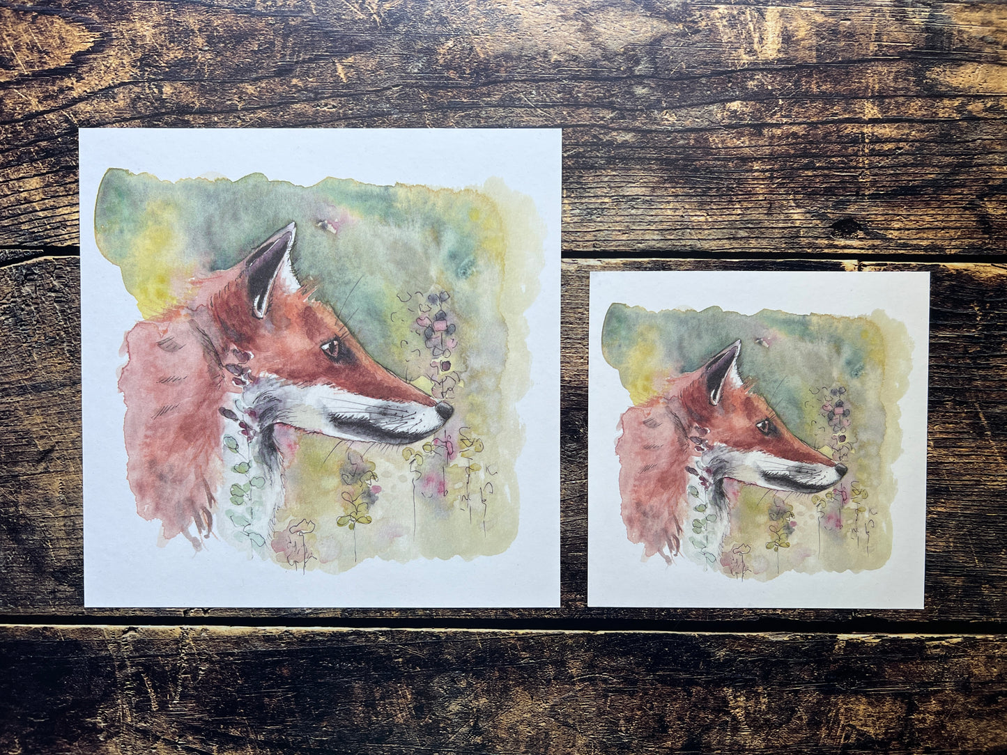 Two watercolor paintings of foxes on a wooden surface