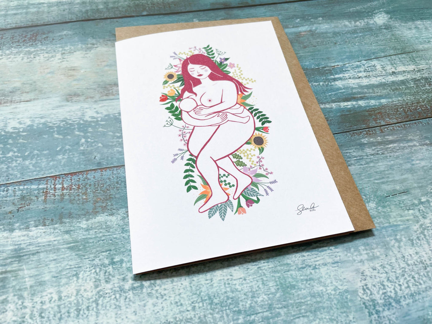 An A6 card of an illustration of a breastfeeding mother laying on a bed of flowers and leaves