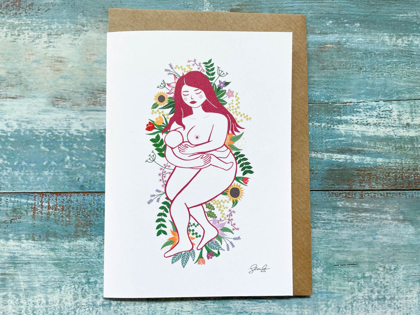 An A6 card of an illustration of a breastfeeding mother laying on a bed of flowers and leaves