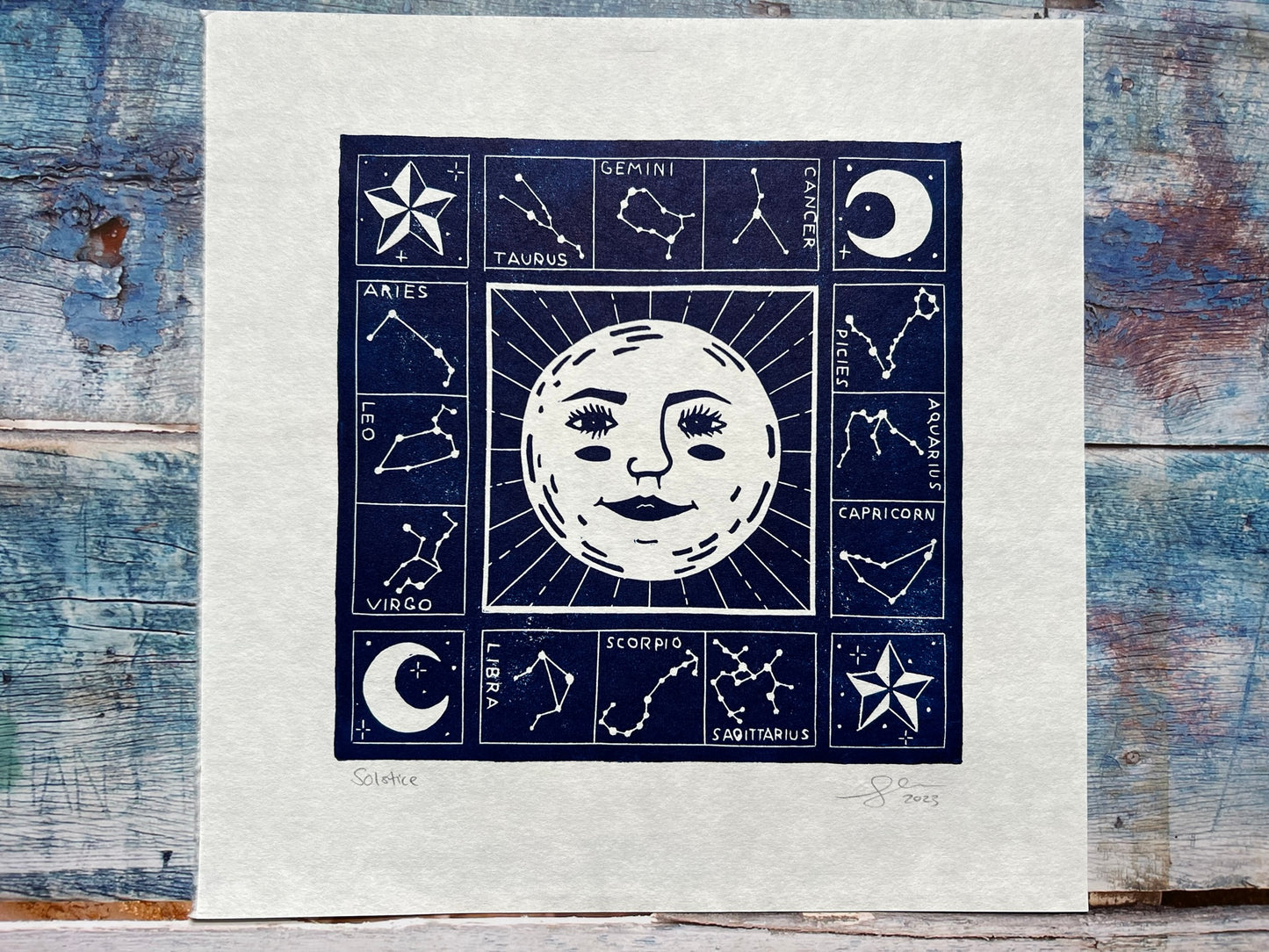 A photo of a dark blue lino print on a blue wooden background. The print is of a sun (with a face) and surrounded by the zodiac constellations.