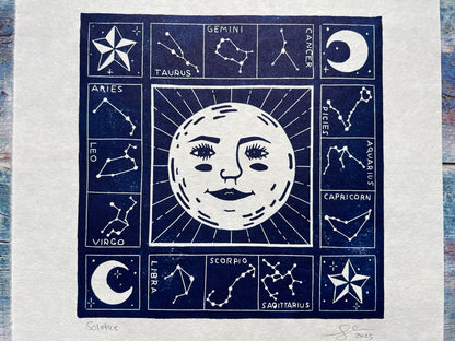 A close up of A photo of a dark blue lino print on a blue wooden background. The print is of a sun (with a face) and surrounded by the zodiac constellations.