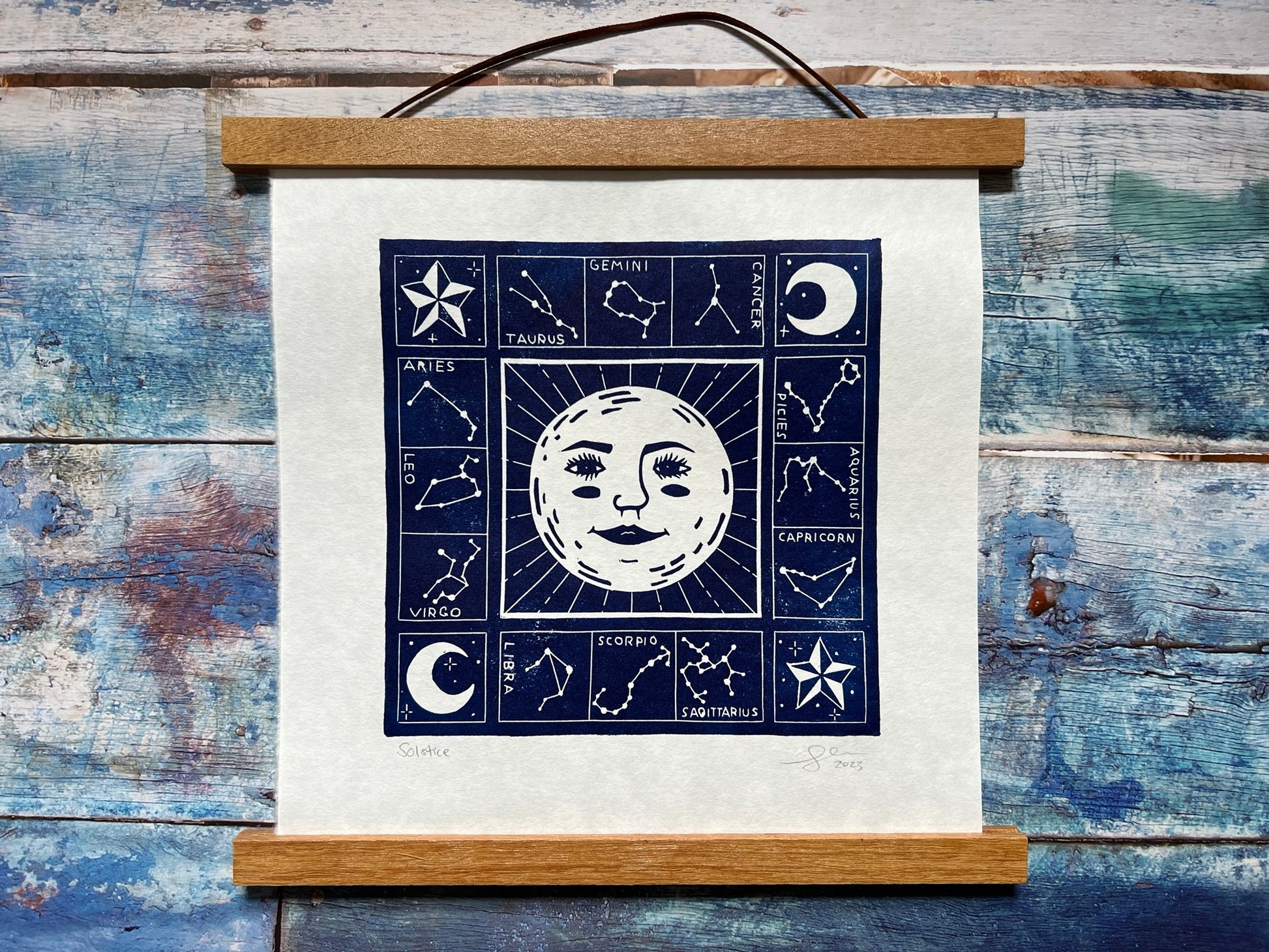 A photo of a dark blue lino print on a blue wooden background. The print is of a sun (with a face) and surrounded by the zodiac constellations. This print is in a magnetic wooden frame.