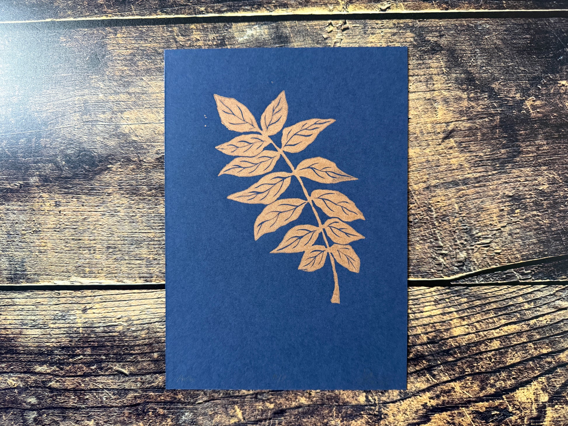 An A5 lino print of an ash tree stem, printed in copper ink on blue paper
