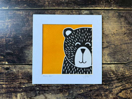 A small square lino print of a black bear with a bright orangey yellow background, mounted