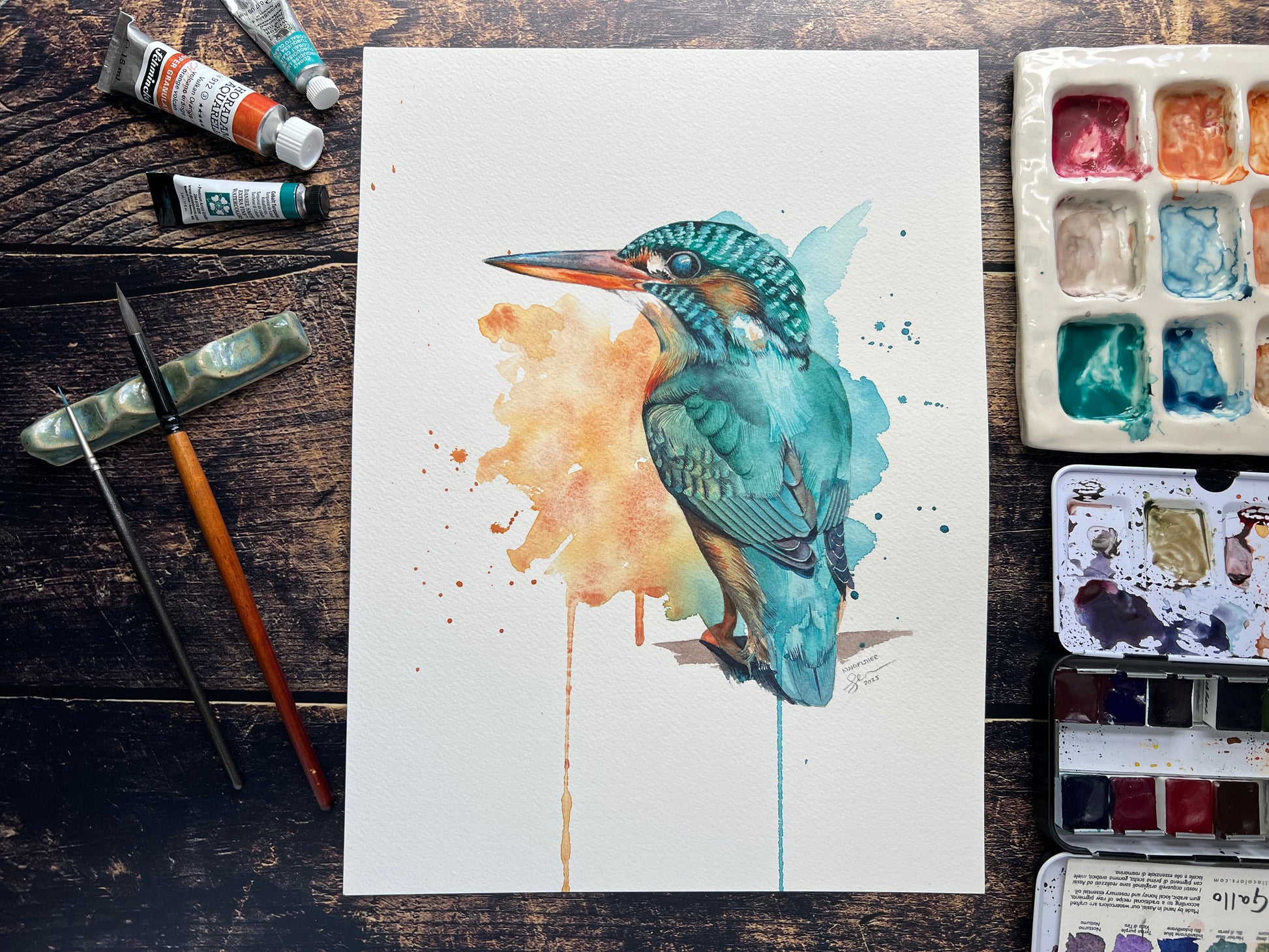 Watercolor painting of a kingfisher on a wooden surface with art supplies.