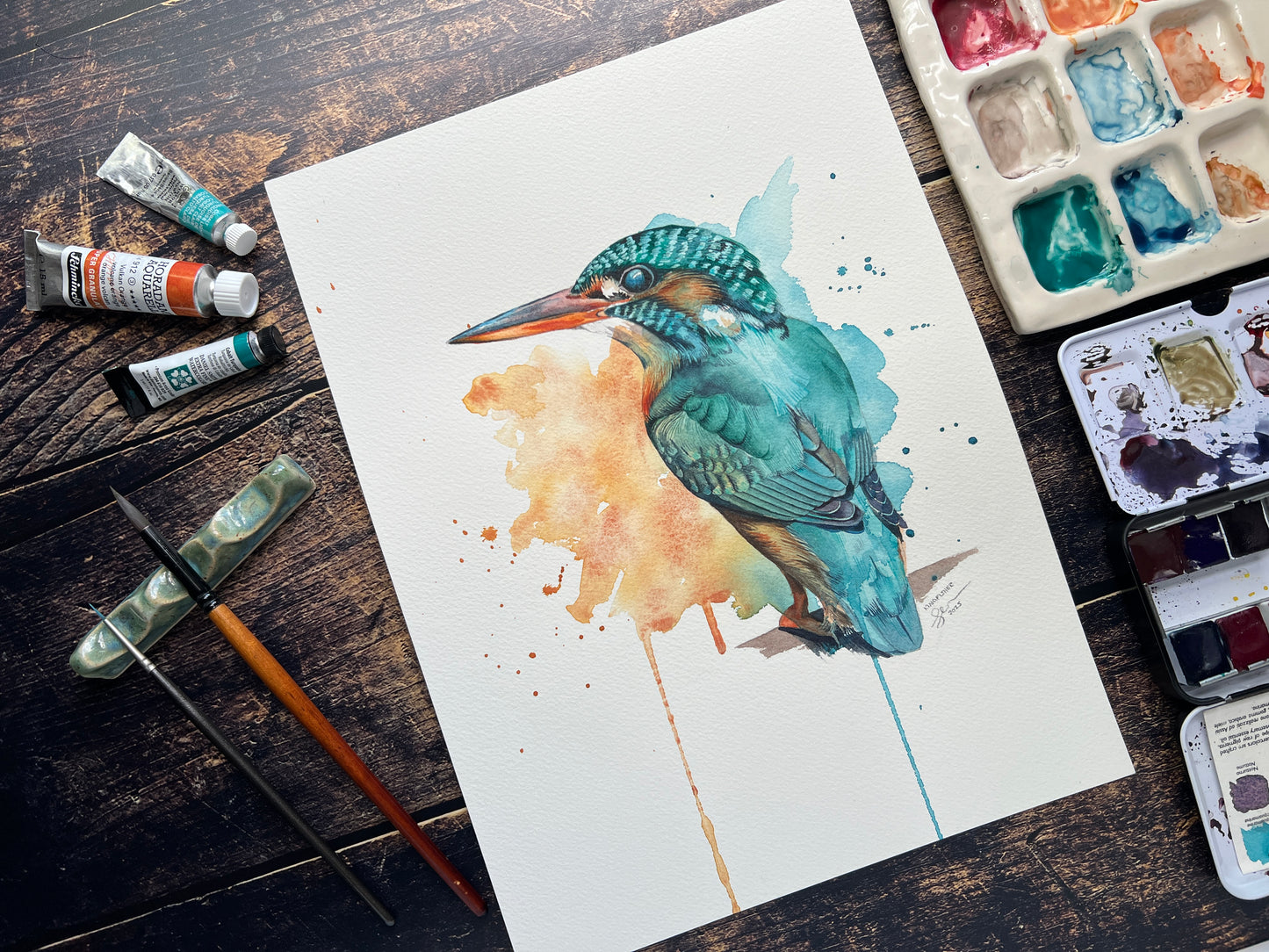 Watercolor painting of a kingfisher on paper with art supplies on a wooden surface