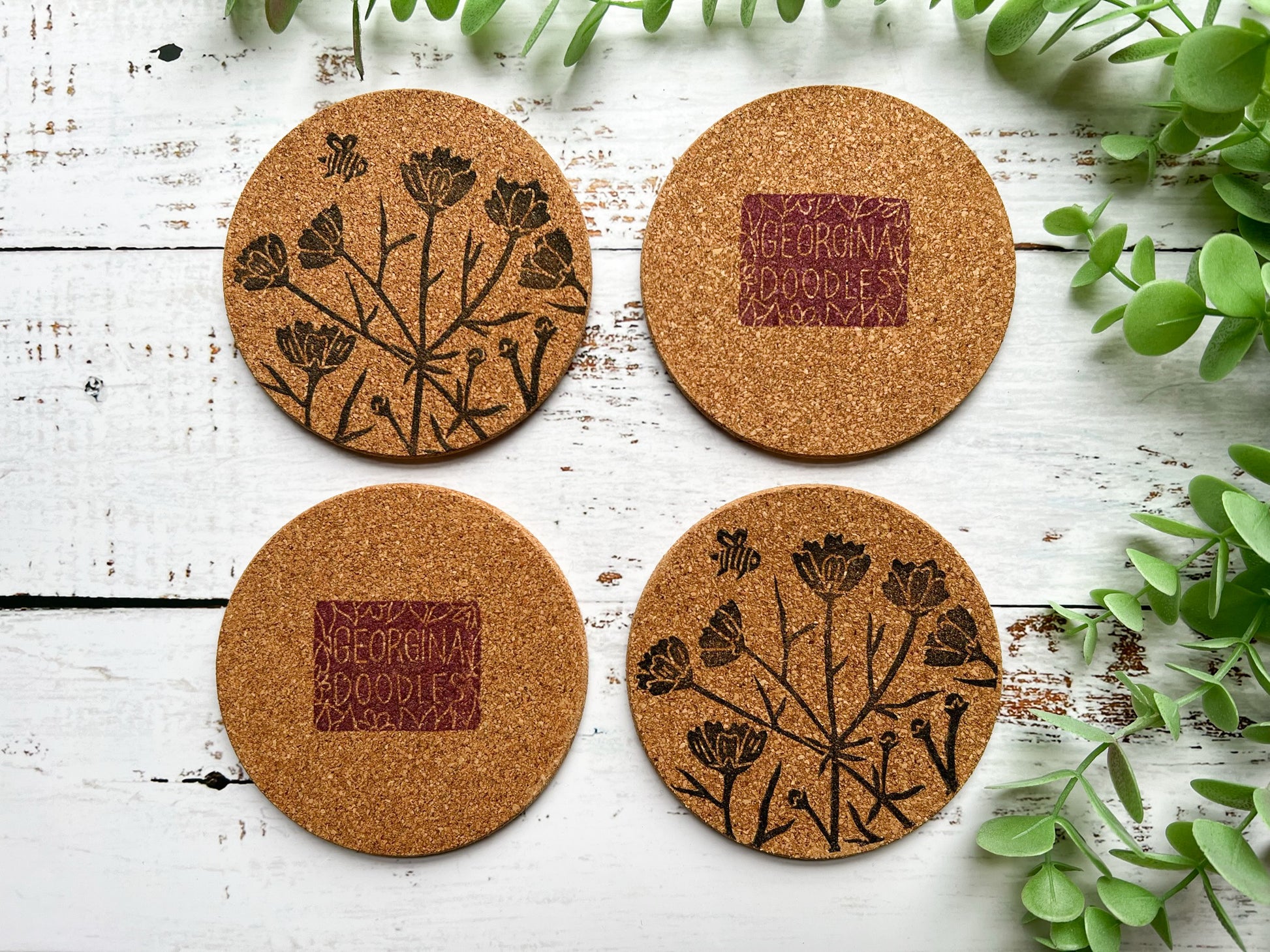 A set of four natural cork coasters with a black floral motif including a bee. Two coasters have been turned over to reveal the underneath and show the logo.