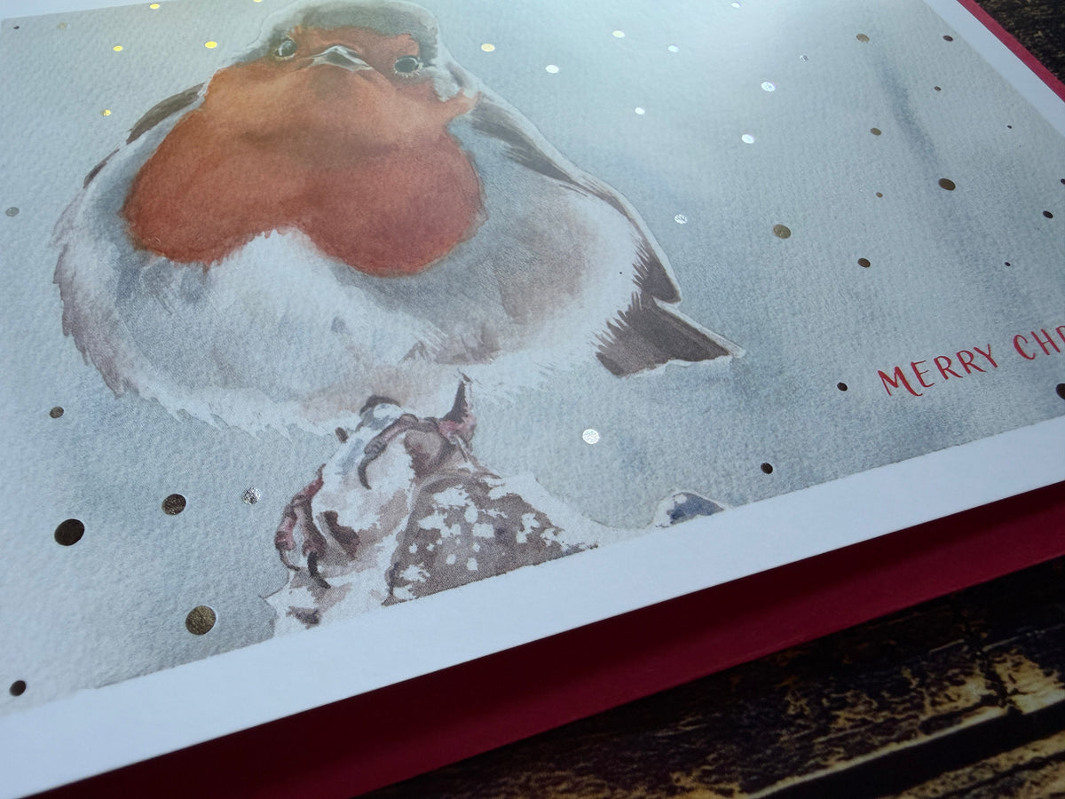 A Christmas card featuring a watercolour painting of a robin, surrounded by silver foil snow.
