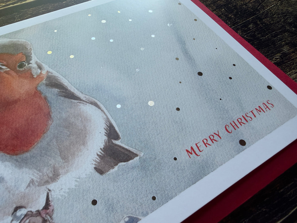 A Christmas card featuring a watercolour painting of a robin, surrounded by silver foil snow.