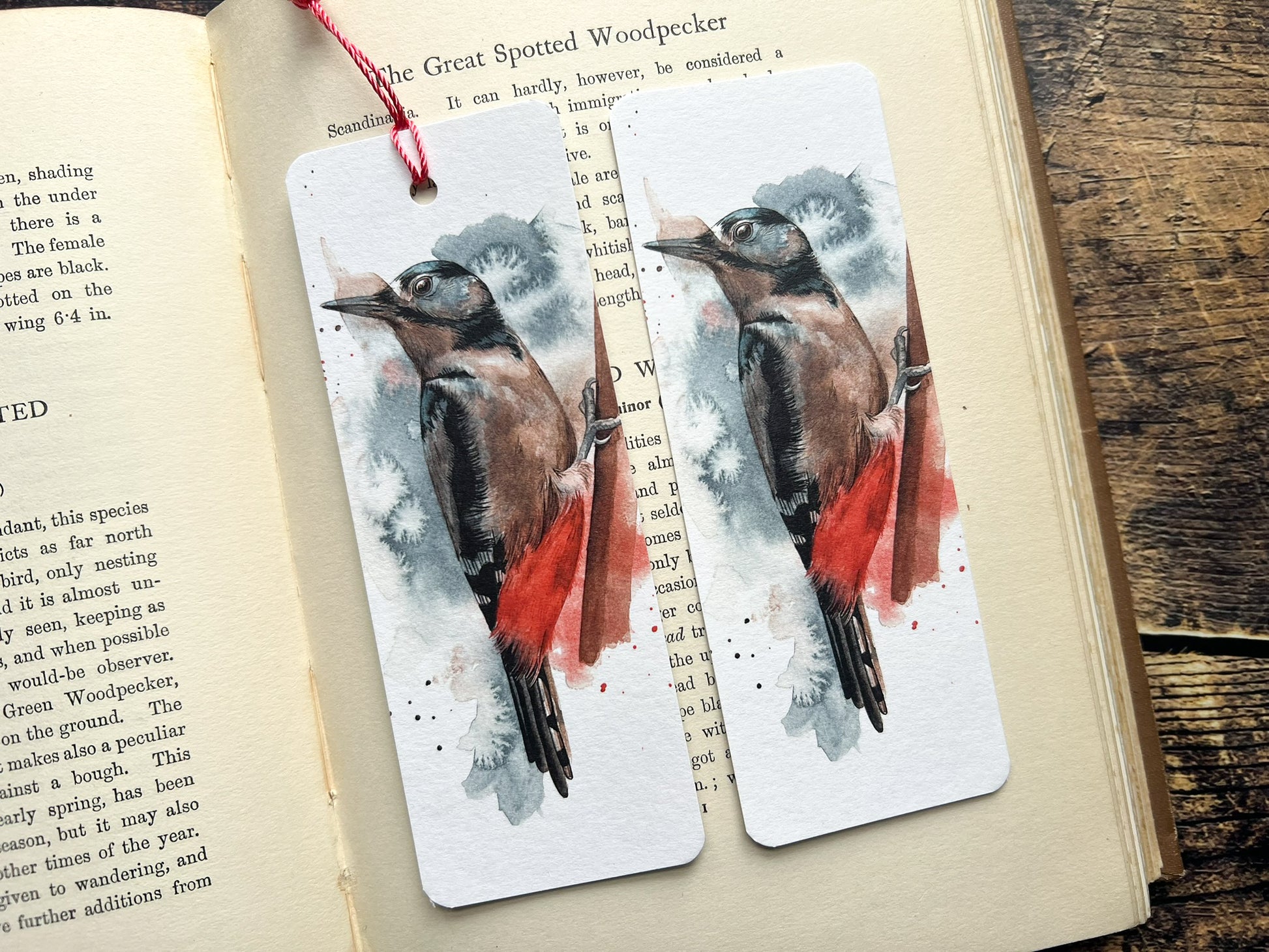 A photo of a bookmark of a great spotted woodpecker watercolour, photographed on an open book. One has a tassel one doesn't.