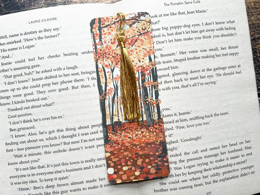 A photo of a bookmark placed on an open book. The bookmark design is an autumnal woodland scene painted in gouache.