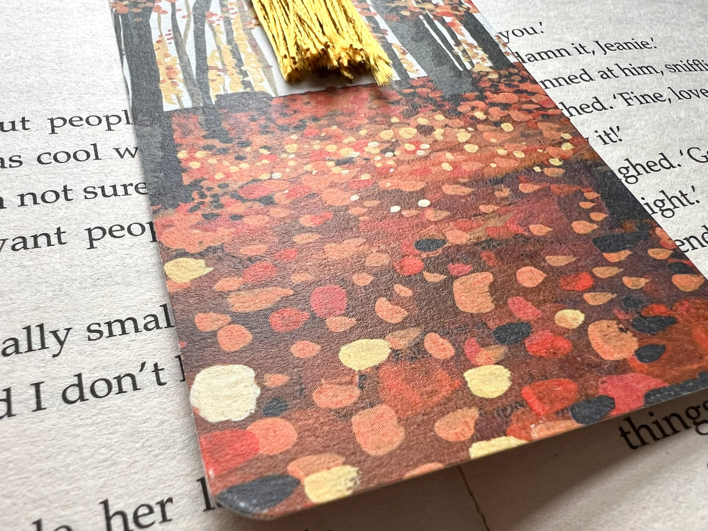 A photo of a bookmark placed on an open book. The bookmark design is an autumnal woodland scene painted in gouache.