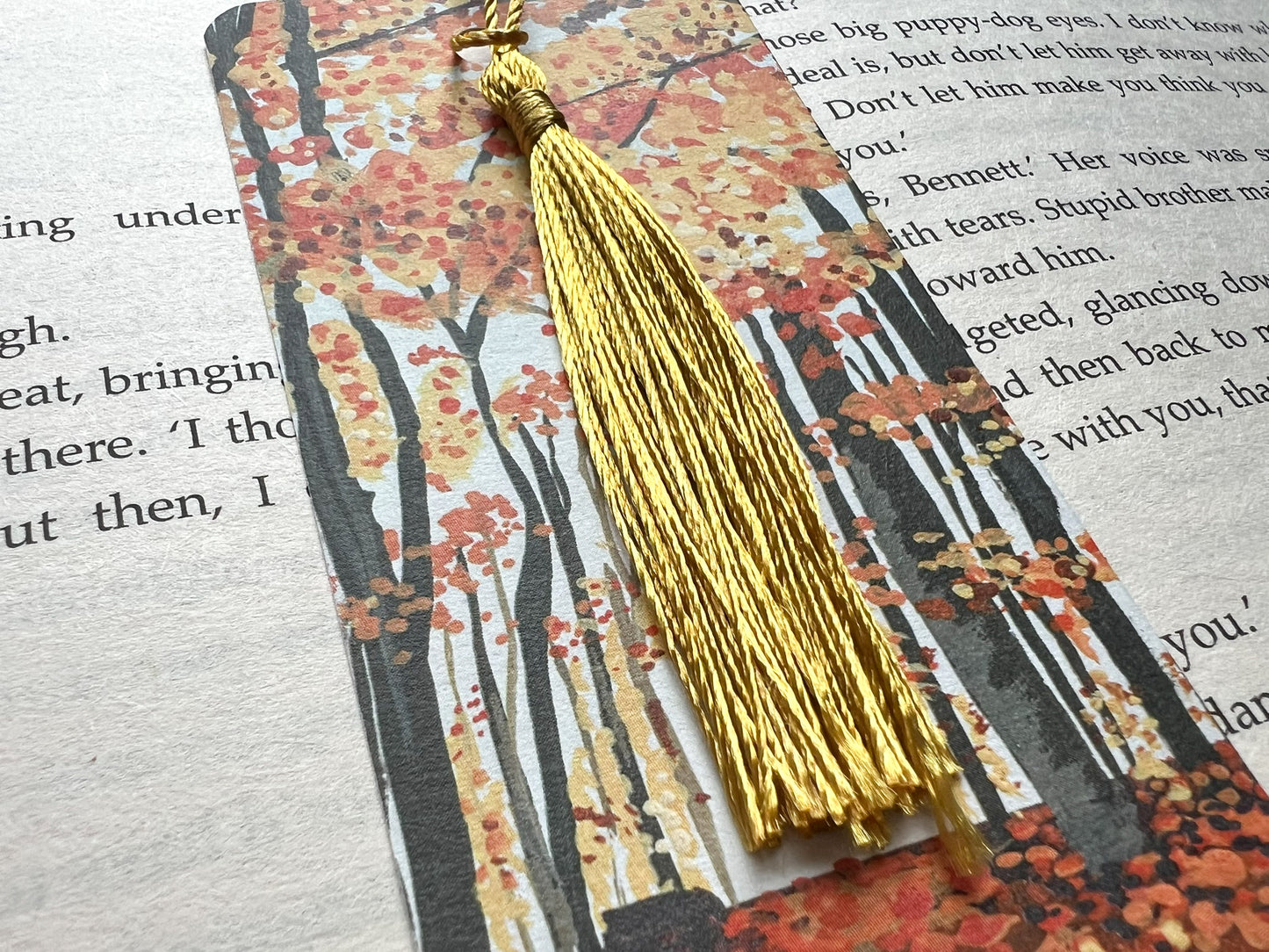 A photo of a bookmark placed on an open book. The bookmark design is an autumnal woodland scene painted in gouache.