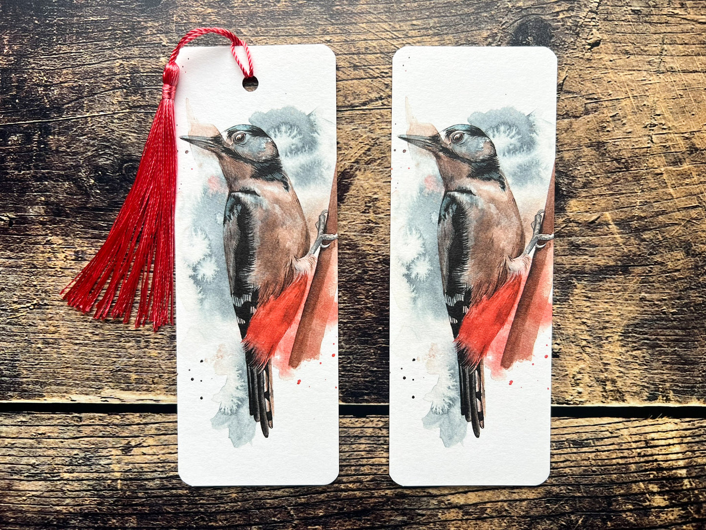 A photo of a bookmark of a great spotted woodpecker watercolour, one with a red tassel and one without.