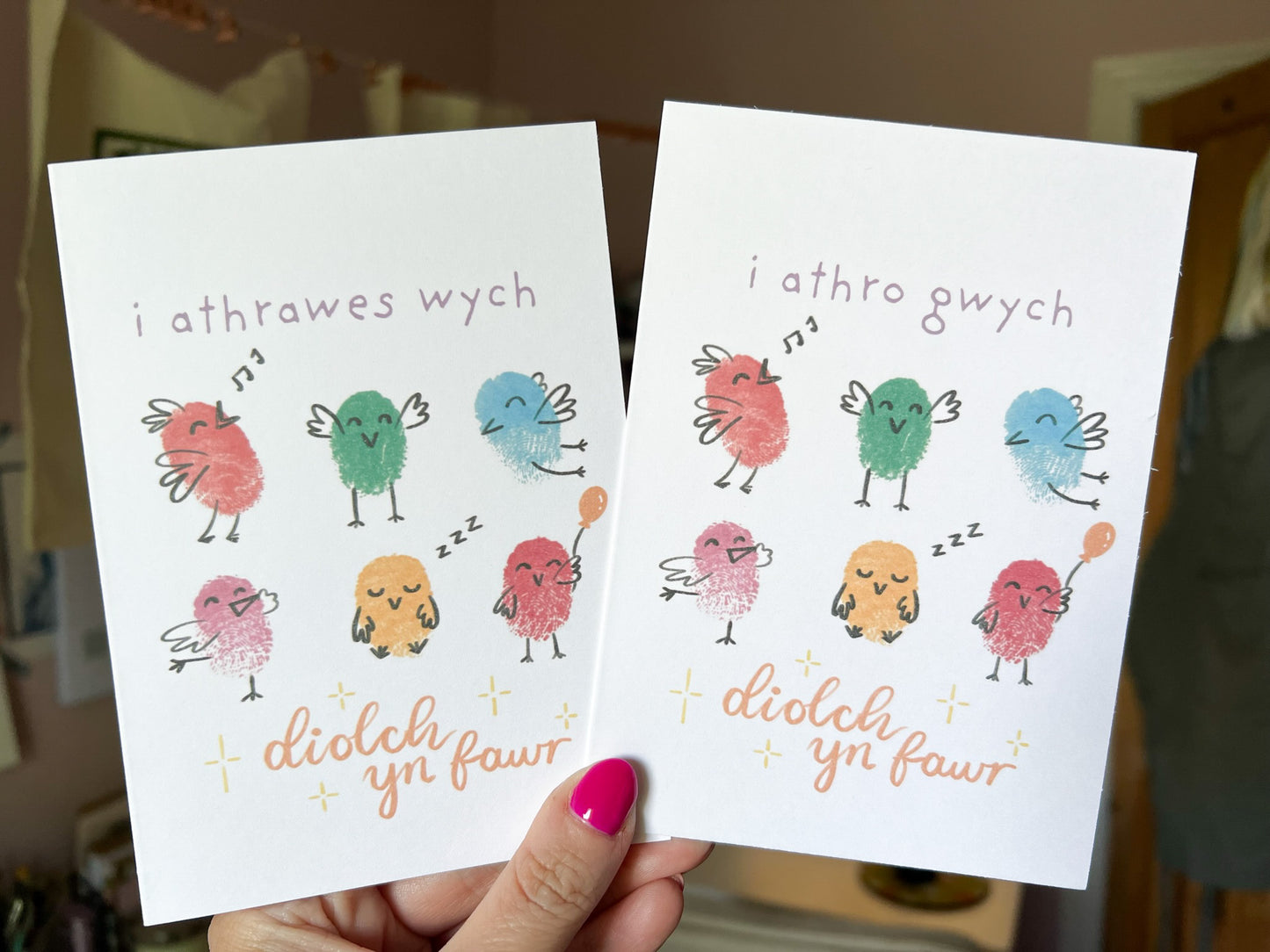 A hand holding two Welsh teacher thank you cards, one says 'i athrawes wych, diolch yn fawr' and the other 'i athro gwych, diolch yn fawr'. Both have six little fingerprinted birds on.