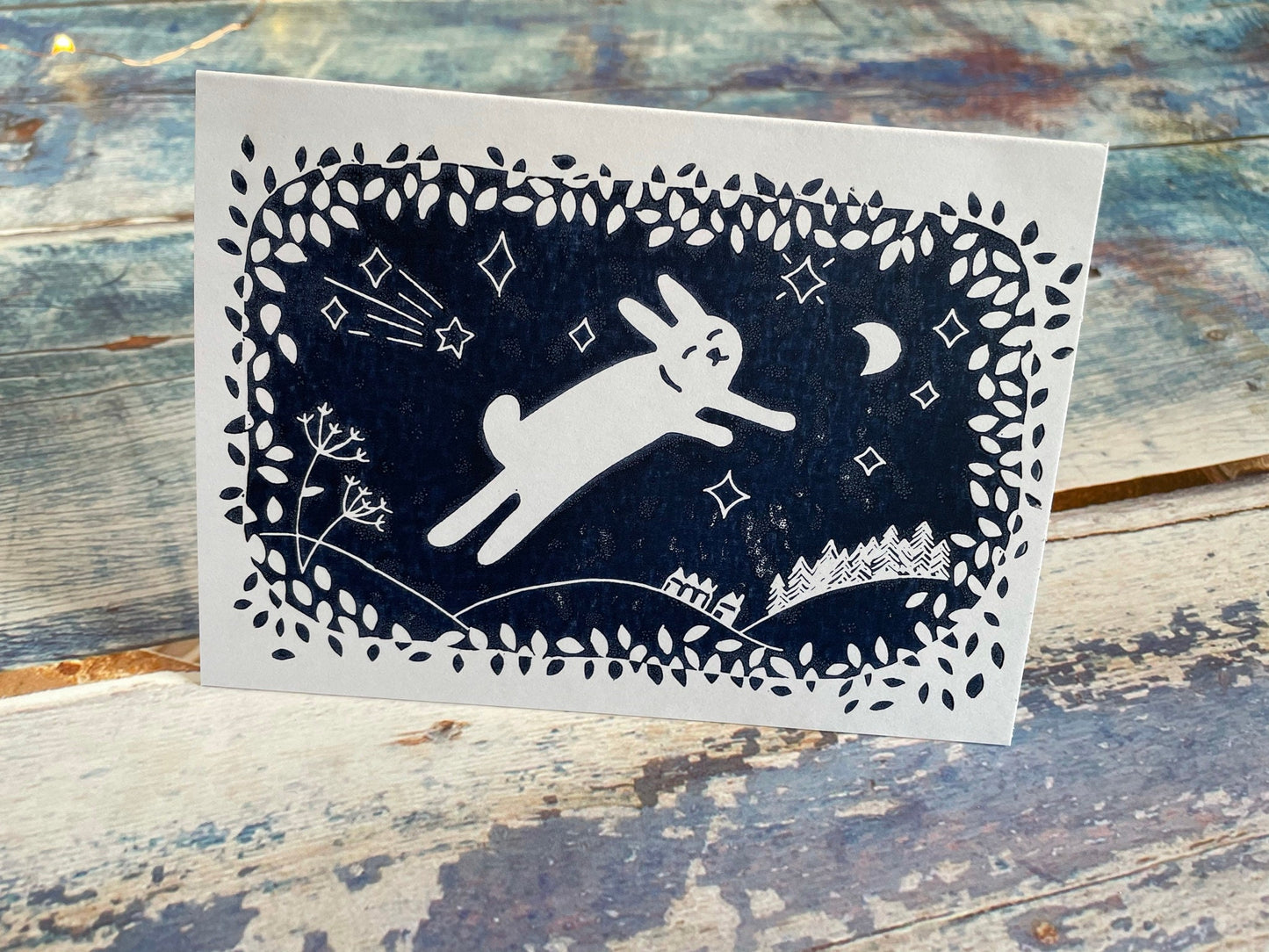 A navy blue lino print of a cute rabbit leaping through the air while surrounded by leaves, moon and stars