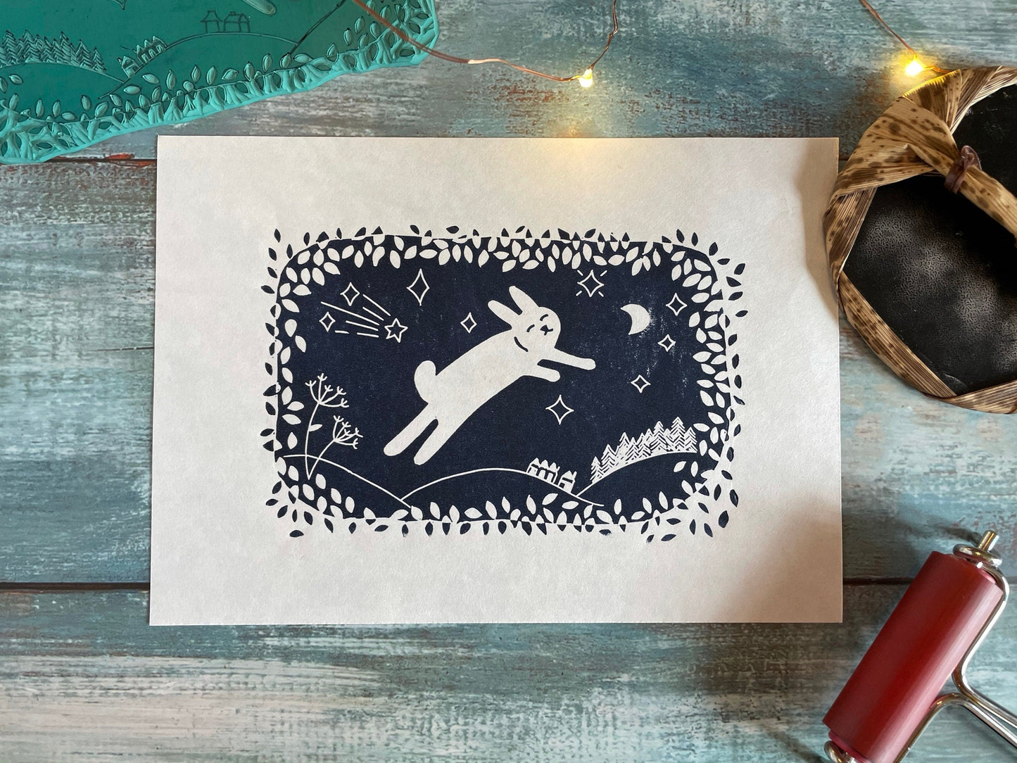 A navy blue lino print of a cute rabbit leaping through the air while surrounded by leaves, moon and stars