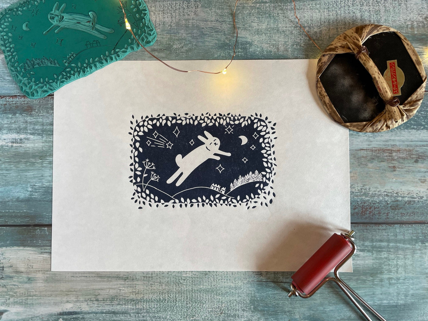 A navy blue lino print of a cute rabbit leaping through the air while surrounded by leaves, moon and stars