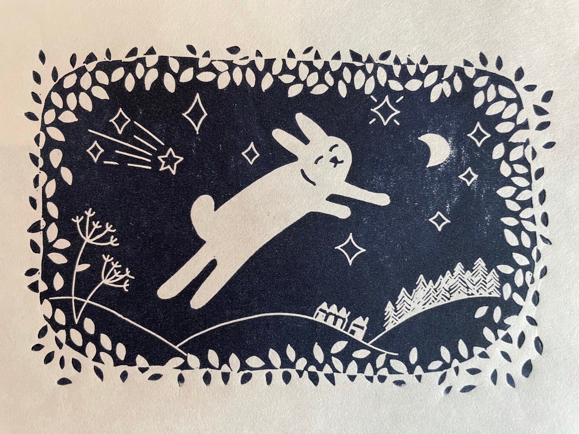 A navy blue lino print of a cute rabbit leaping through the air while surrounded by leaves, moon and stars