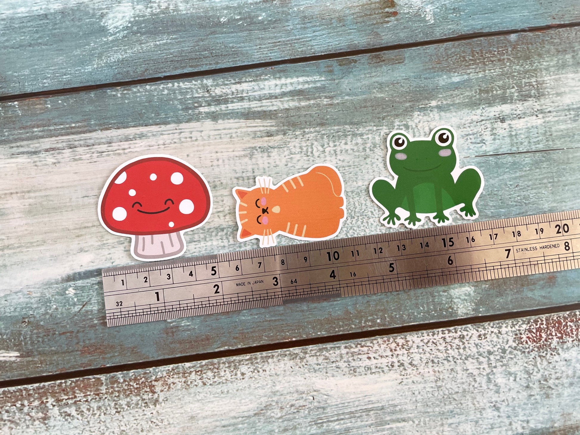 A picture of three 5cm cute stickers, there's a mushroom, a ginger cat and a green frog all with cute faces