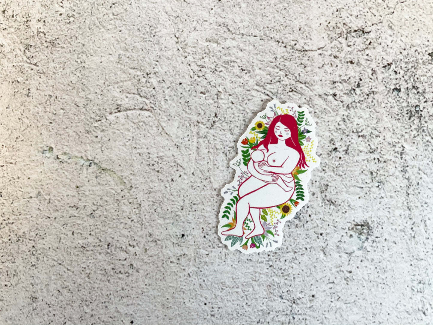 A 5cm sticker of digital art of a naked mother breastfeeding her baby while laying on a bed of flowers and leaves
