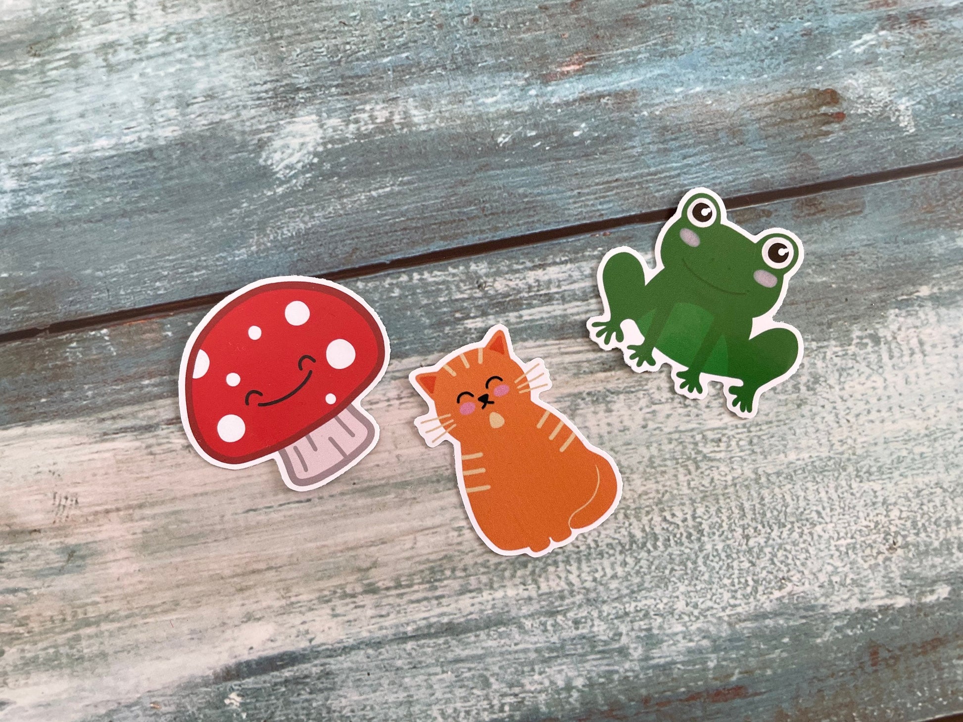 A picture of three 5cm cute stickers, there's a mushroom, a ginger cat and a green frog all with cute faces