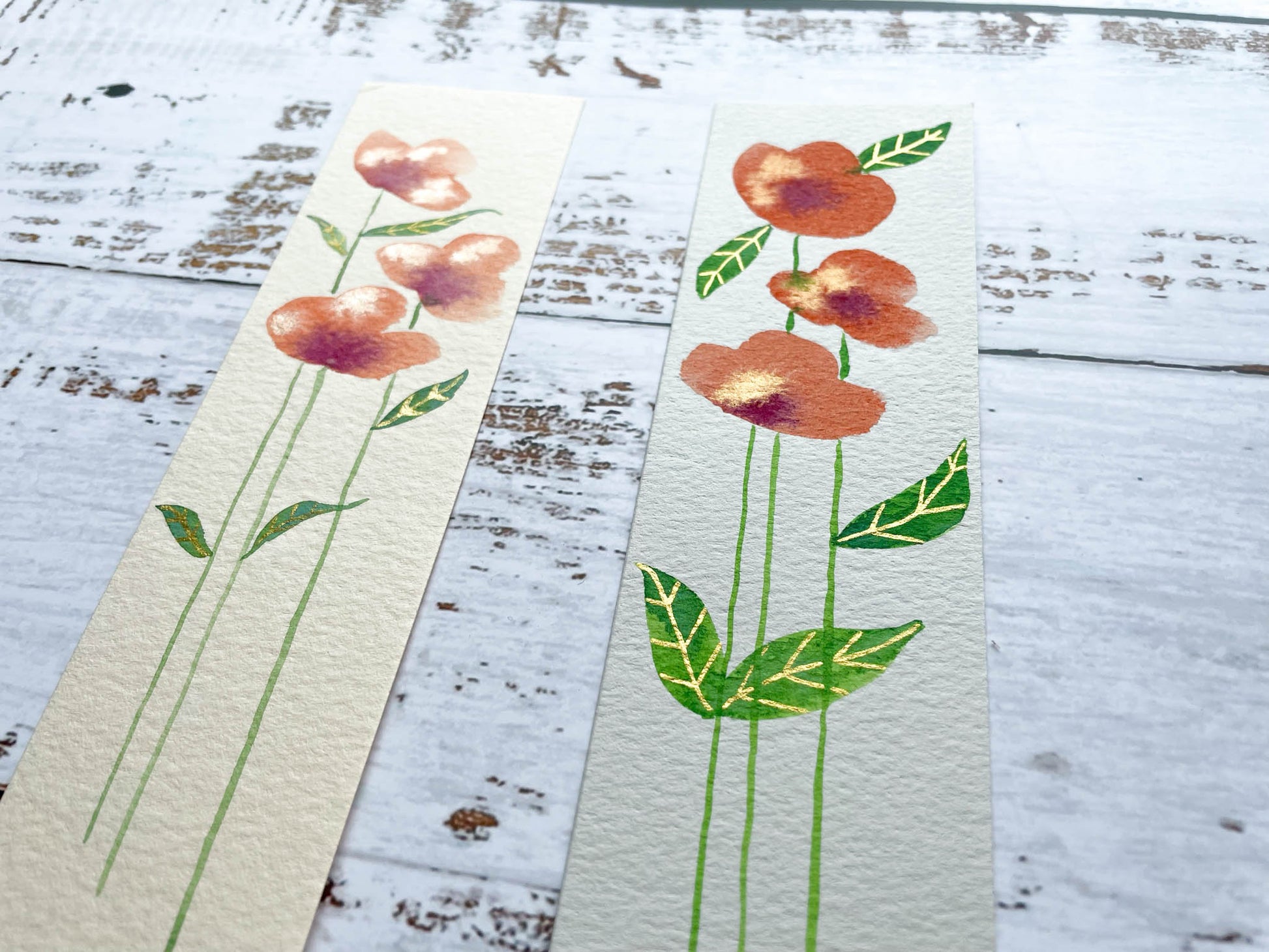 A photo of two hand painted watercolour bookmarks of orange flowers with gold embellishments