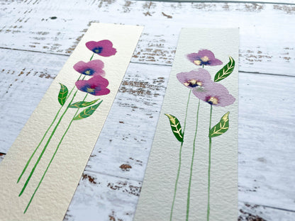 A photo of two hand painted watercolour bookmarks of purple flowers with gold embellishments