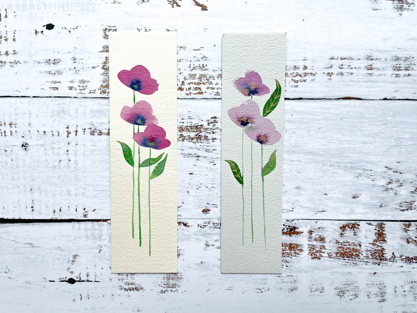 A photo of two hand painted watercolour bookmarks of purple flowers with gold embellishments