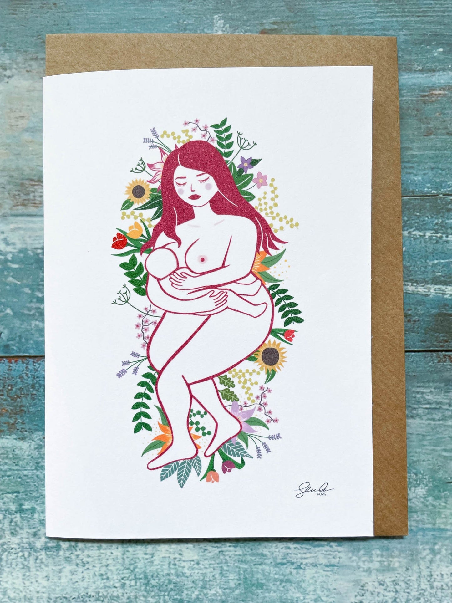 An A6 card of an illustration of a breastfeeding mother laying on a bed of flowers and leaves