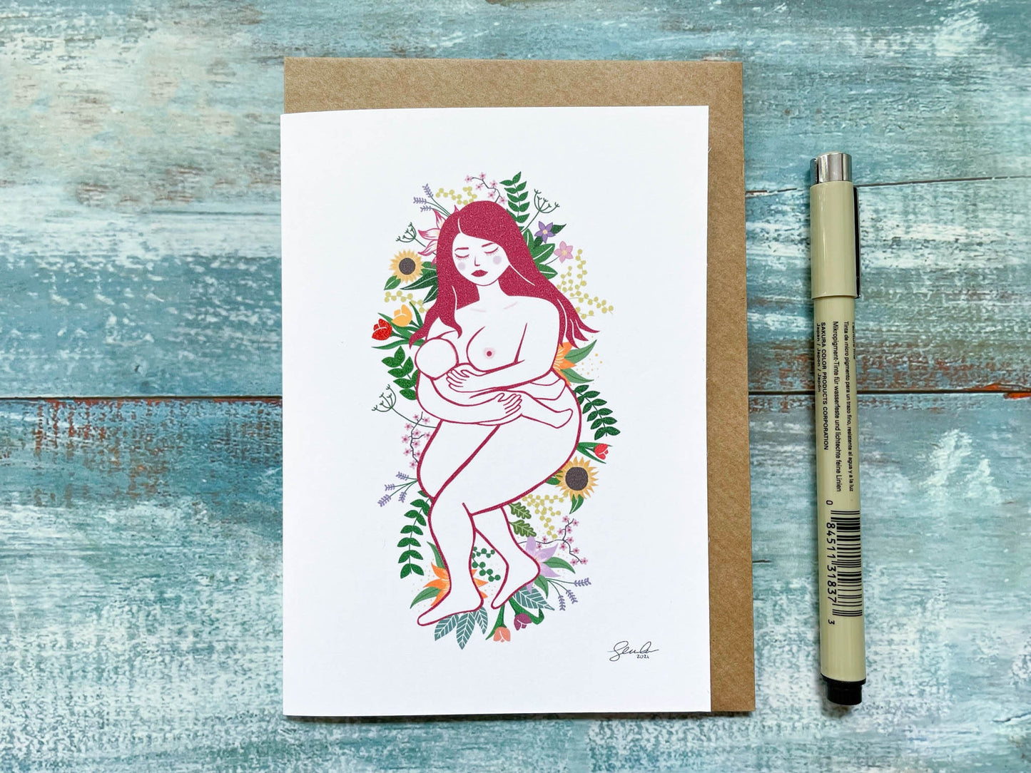 An A6 card of an illustration of a breastfeeding mother laying on a bed of flowers and leaves