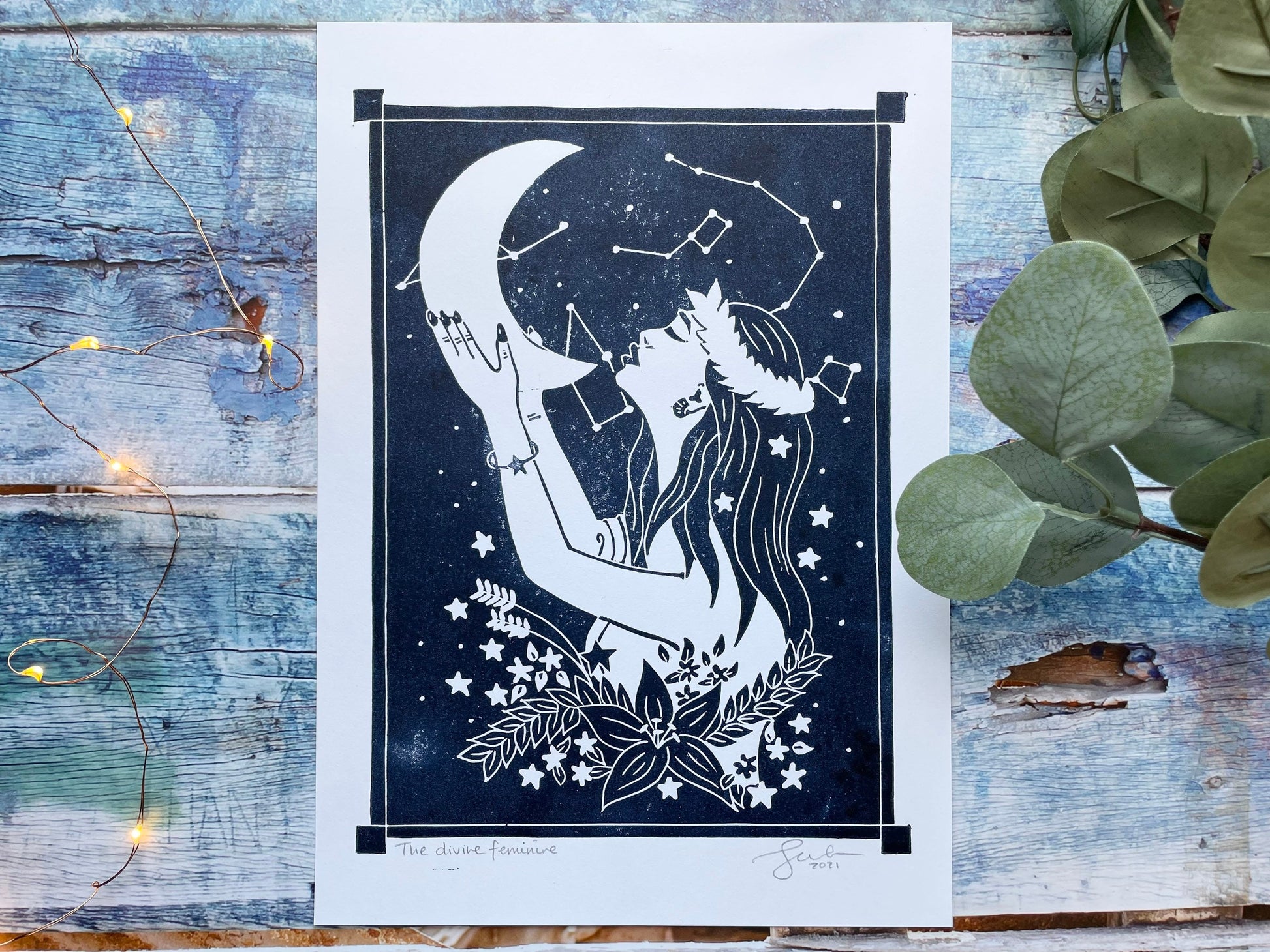 An A4 lino print of a women holding the moon up to her face with her hands, she's surrounded by stars and flowers