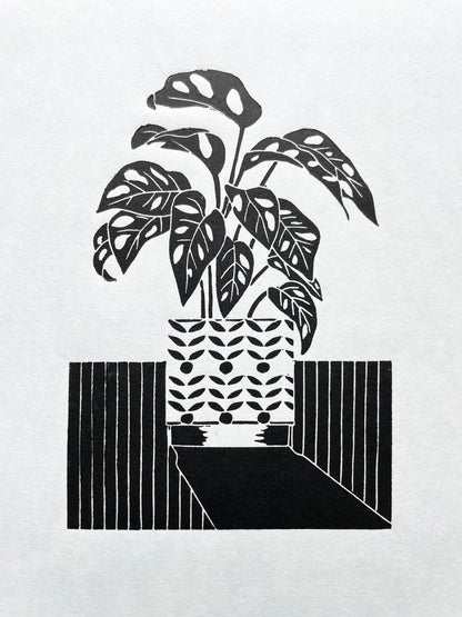 A black lino print of a monstera monkey plant in a patterned plant pot with a long shadow
