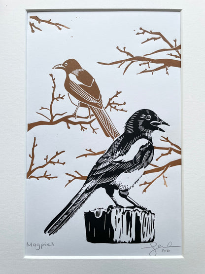 A mounted lino print of two magpies, the one in the foreground is black and the one in the background is brown