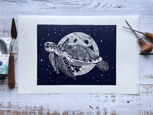 A dark blue lino print of a sea turtle in front of the moon and surrounded by stars, it's printed on paper that's almost A3