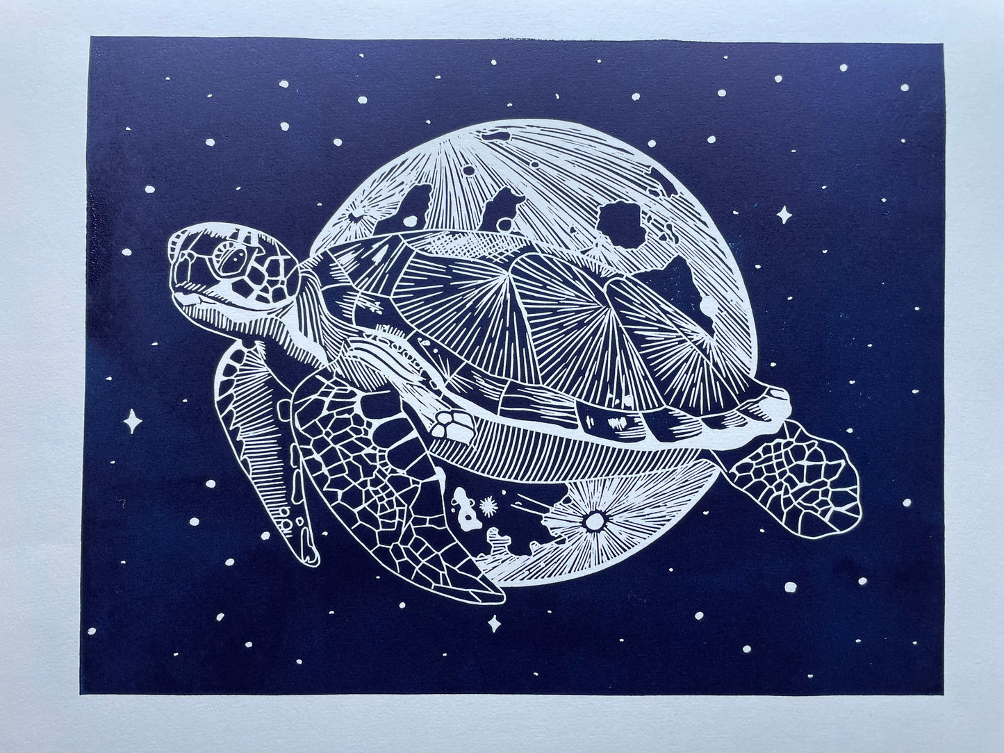 A dark blue lino print of a sea turtle in front of the moon and surrounded by stars, it's printed on paper that's almost A3