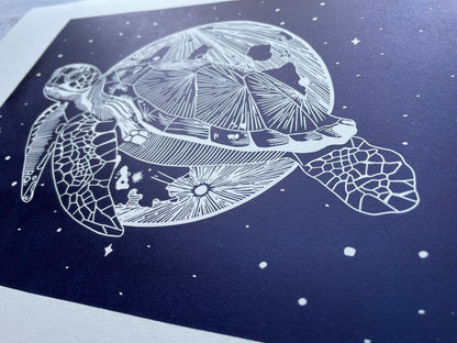 A dark blue lino print of a sea turtle in front of the moon and surrounded by stars, it's printed on paper that's almost A3