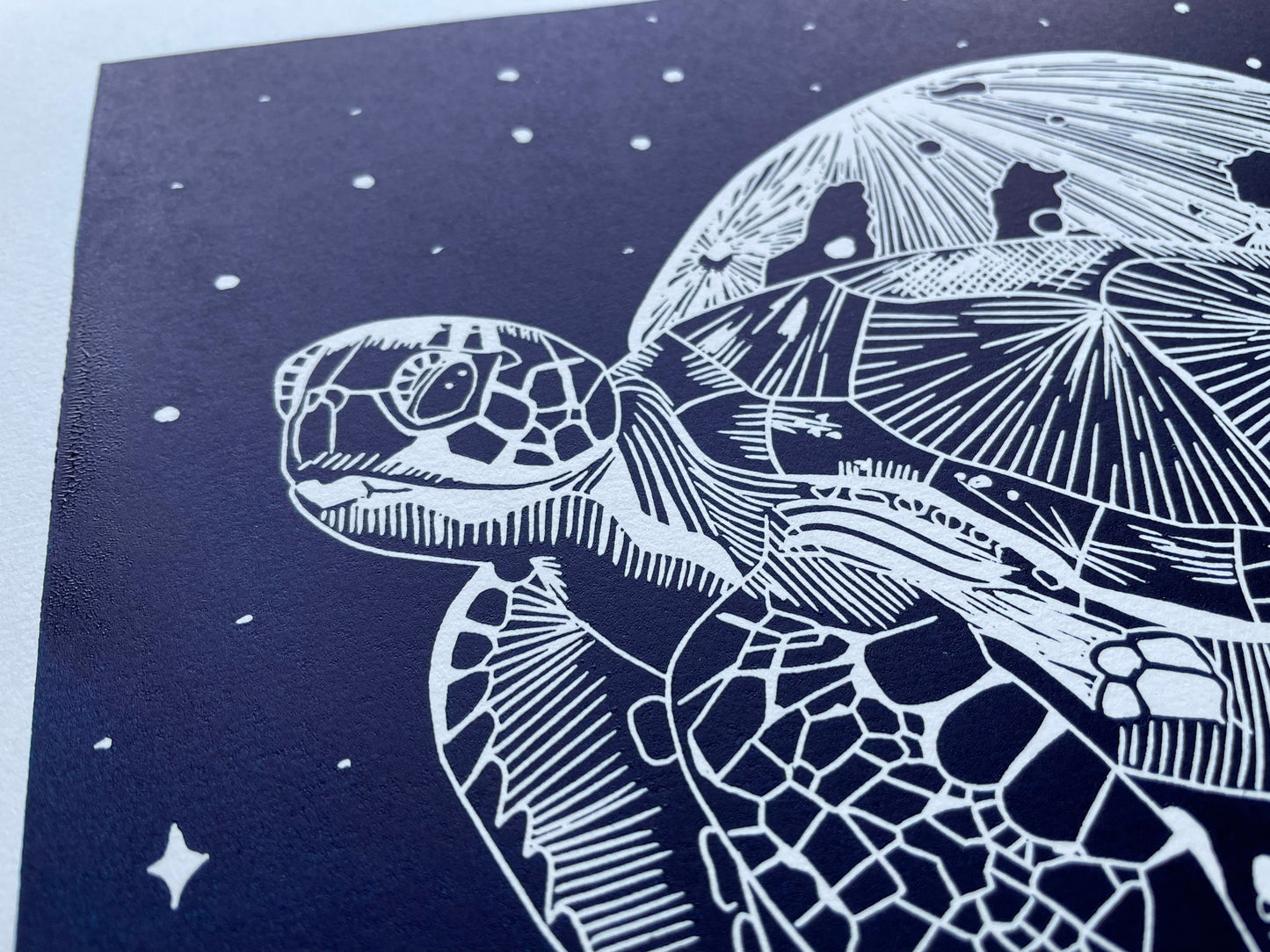 A dark blue lino print of a sea turtle in front of the moon and surrounded by stars, it's printed on paper that's almost A3