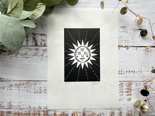 A lino print of a sun with a face and rays coming away from her.
