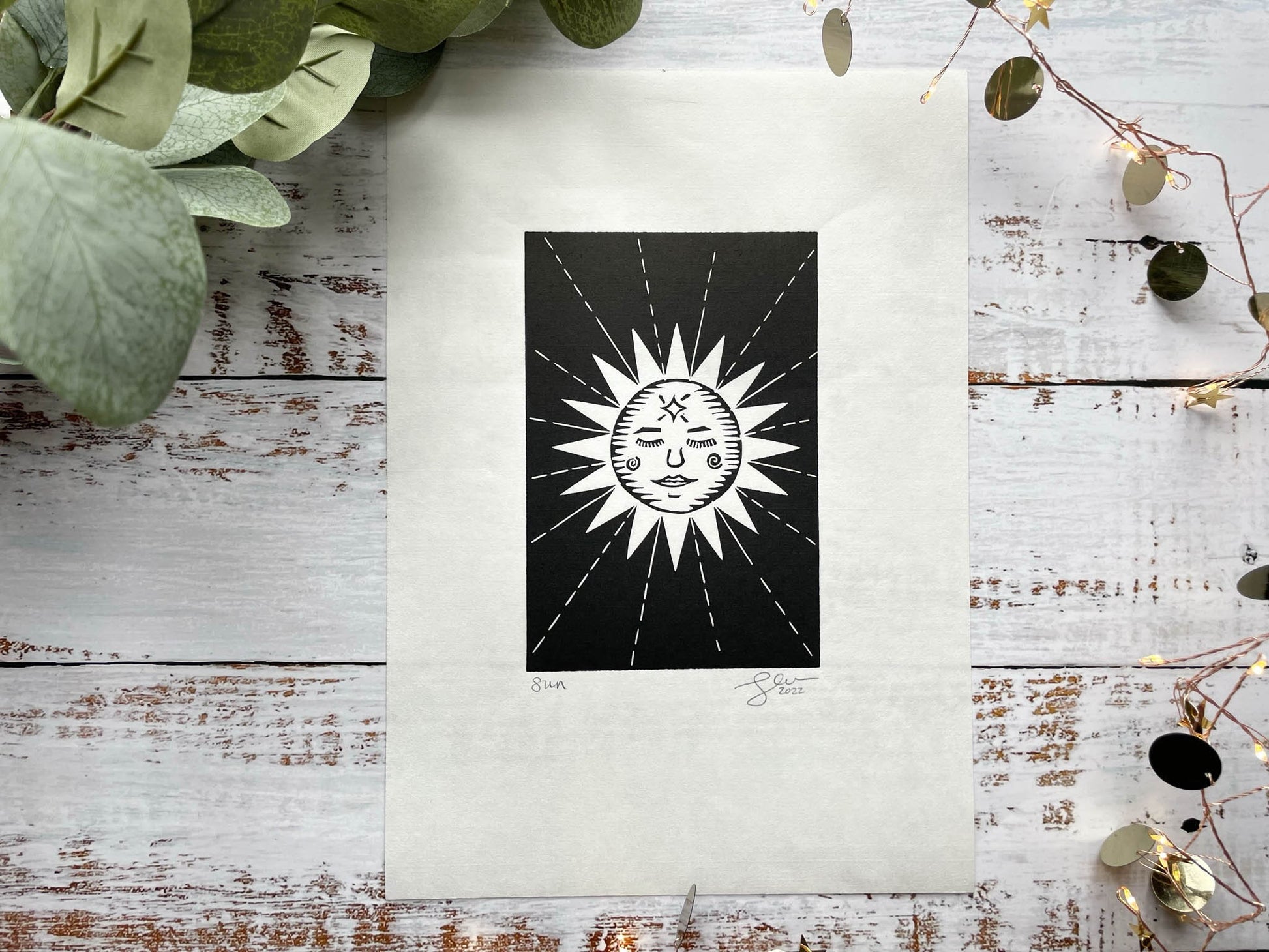 A lino print of a sun with a face and rays coming away from her.