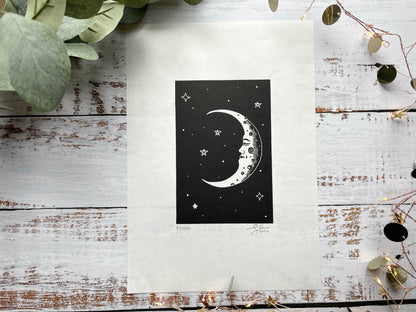 A black lino print of a crescent moon with a face