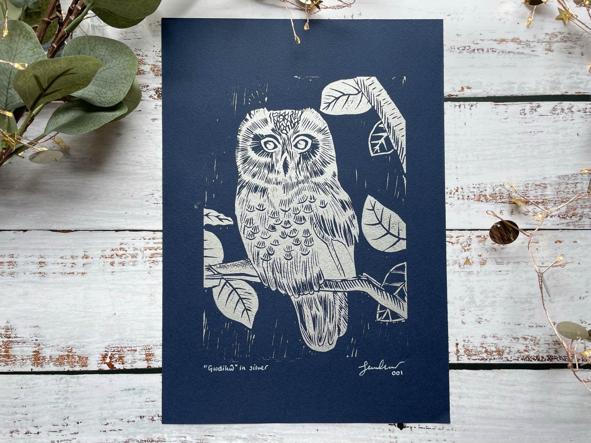 A lino print of an owl sitting in a tree in silver on navy blue paper