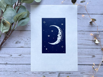 A blue lino print of a crescent moon with a face