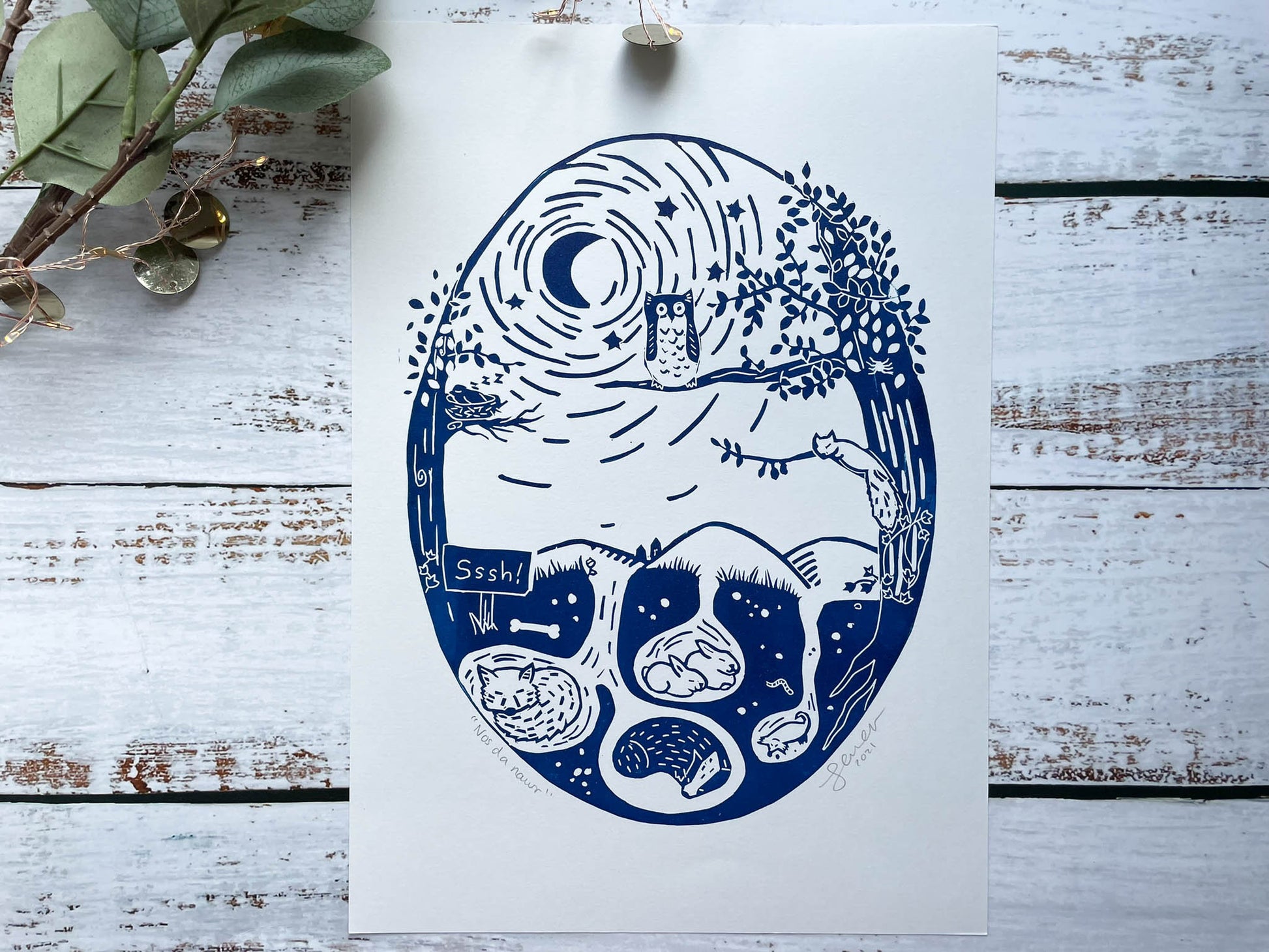 An oval lino print of an owl in a tree watching over the woodland animals while they sleep in blue