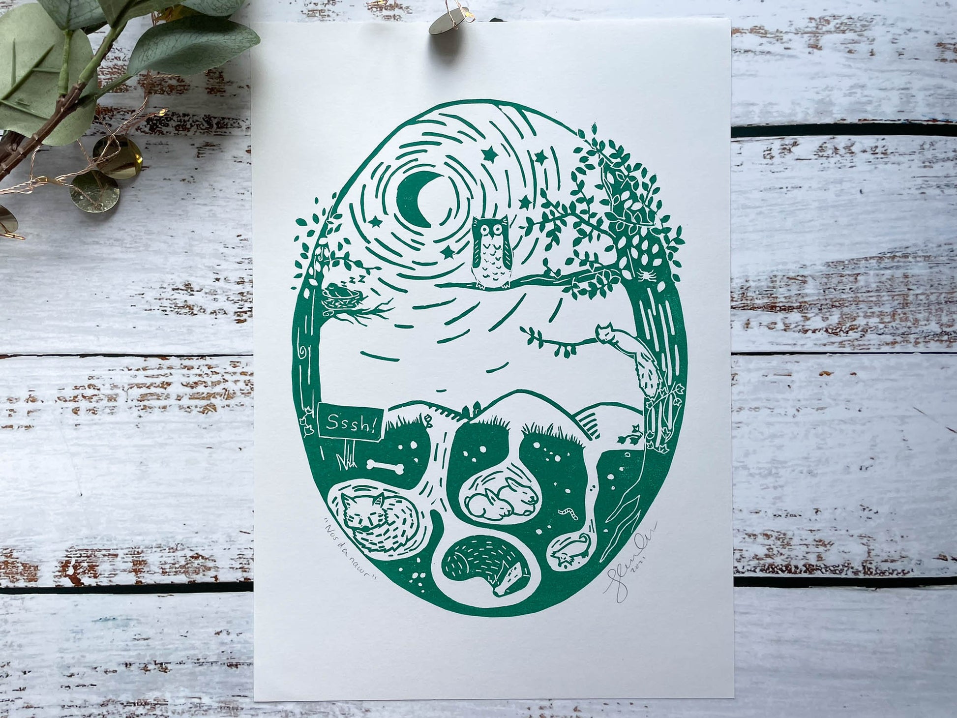 An oval lino print of an owl in a tree watching over the woodland animals while they sleep in green