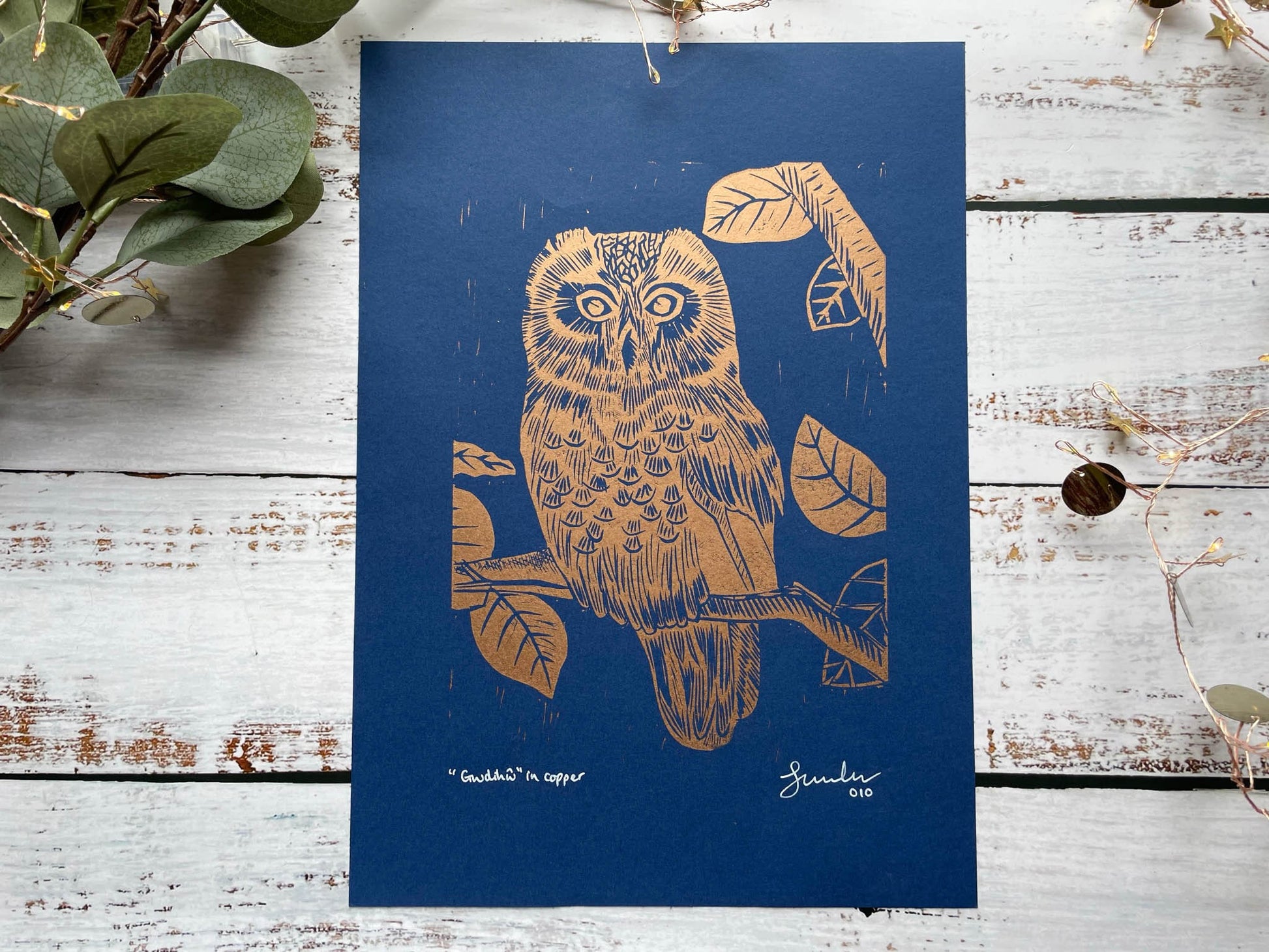 A lino print of an owl sitting in a tree in copper on blue paper