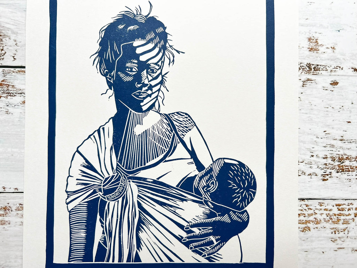 A blue lino print of a mother breastfeeding her baby in a ring sling with sunlight across her face through the blinds.